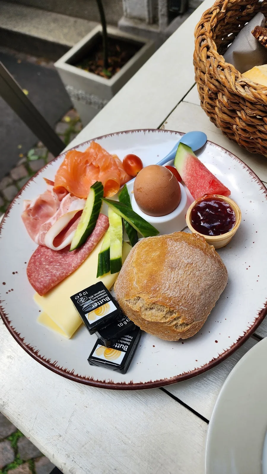 Frühstücksteller mit Lachs, Schinken, Salami, Käse, gekochtem Ei, Gurke, Melone, Marmelade und einem Rundstück.