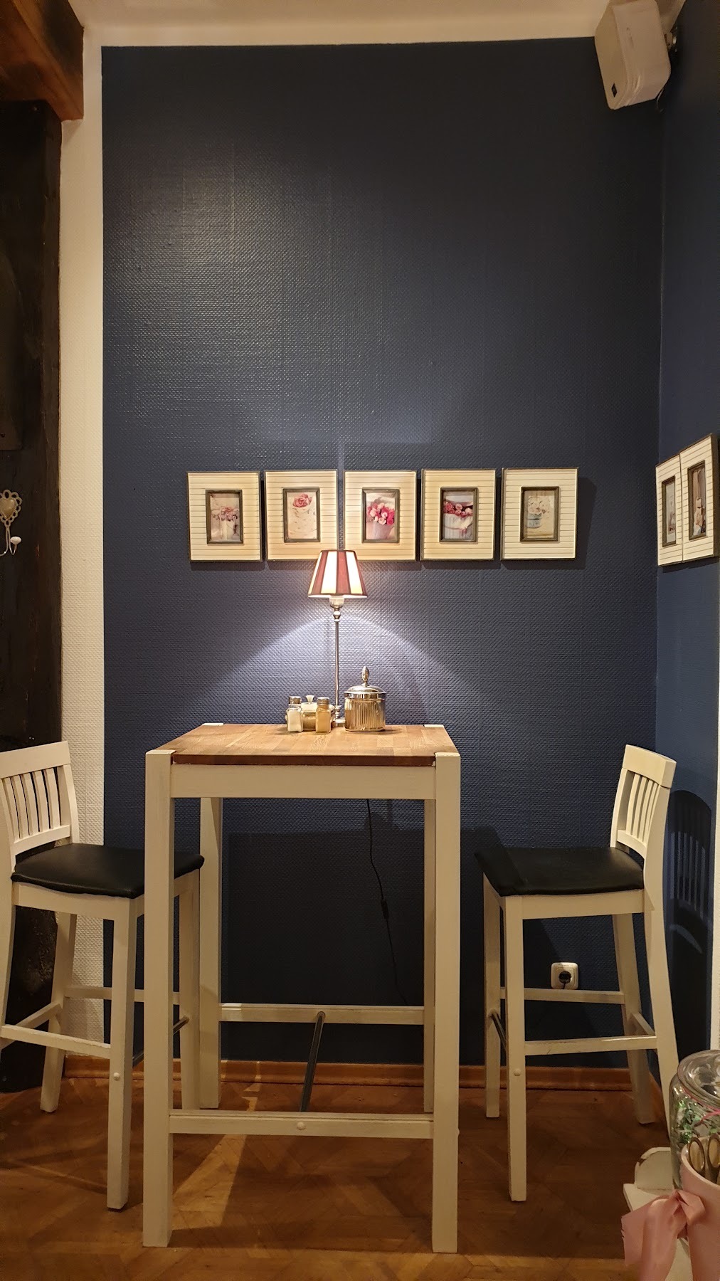 Hoher Tisch vor dunkelblauer Wand, darüber eine Reihe kleiner gerahmter Blumenbilder und eine Tischlampe mit gestreiftem Schirm.