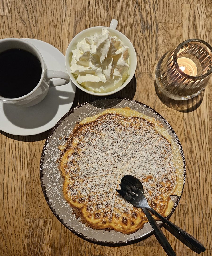 Waffel mit Puderzucker, eine Tasse schwarzer Kaffee, ein Schälchen Sahne und ein brennendes Teelicht auf einem Holztisch.